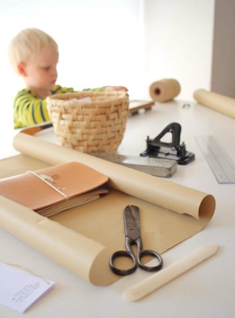 Ein Kleinkind im Hintergrund eines hellen Arbeitstisches mit Lederprodukten, Packpapier und einer alten Handwerksschere bei Lederlotta. Lederlotta Anna Lewis